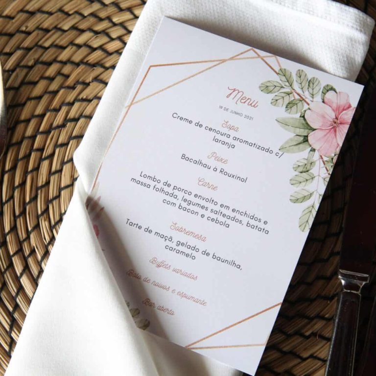 Menu de casamento com lista de pratos, decorado com flores e folhas, colocado sobre uma toalha branca em uma mesa de jantar rústica.
