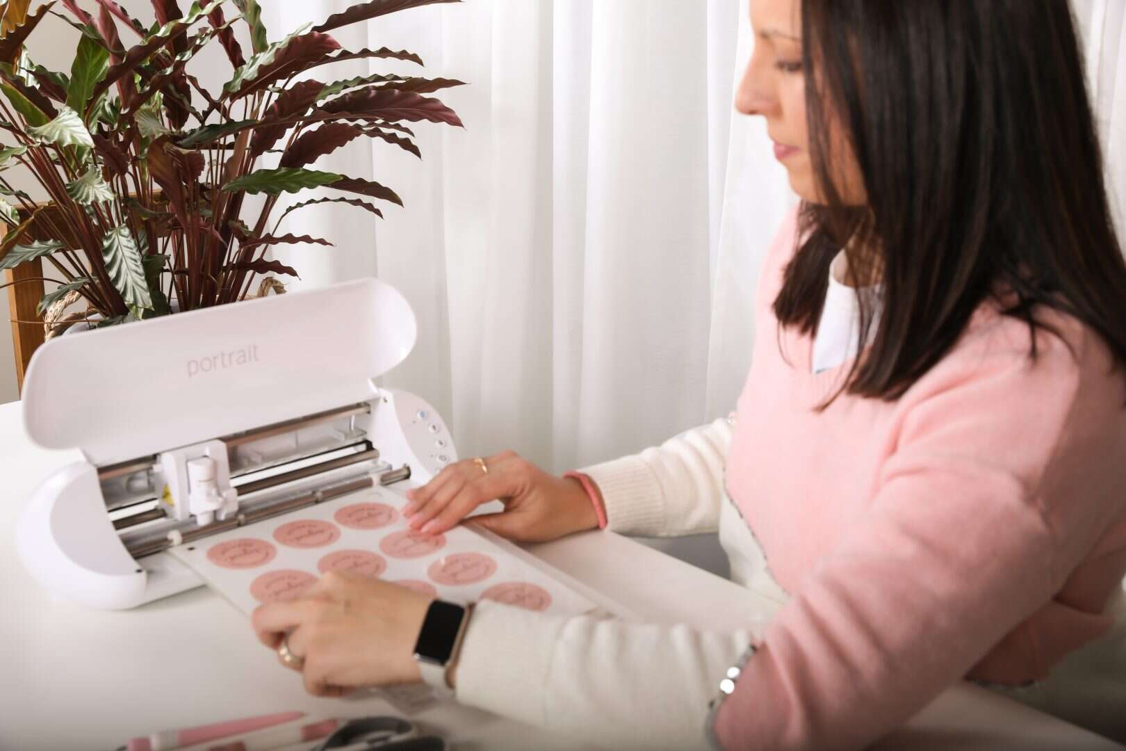 Mulher usando uma impressora de rosto para criar maquilhagem detalhada, sentada numa mesa com uma planta ao fundo, em ambiente bem iluminado.