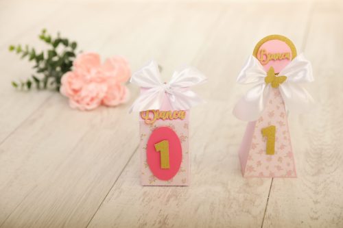 Dois bolos decorativos de primeira aniversário, um com número 1 em rosa e outro com o nome "Diana" em dourado, decorados com fitas de cetim brancas e detalhes delicados.