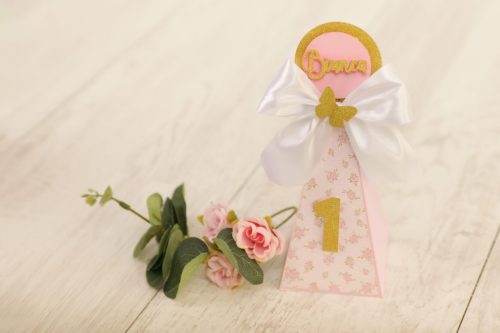 Boneca de papel decorativa com o nome "Bruna" em dourado, uma fita de cetim branca com laço e uma flor de papel rosa, usada como decoração de festa de aniversário de primeira idade.