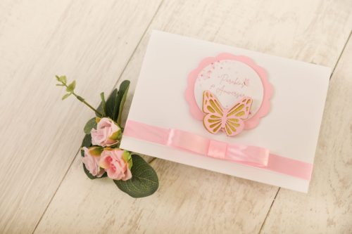 Padrão de cartão de convite com borboleta dourada e laço rosa, decorado com flores rosas, colocado sobre uma superfície de madeira clara.