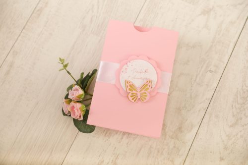 Padrão de cartão de aniversário rosa com borboleta decorativa e mensagem, acompanhado de flores artificiais, colocado sobre uma superfície de madeira clara.