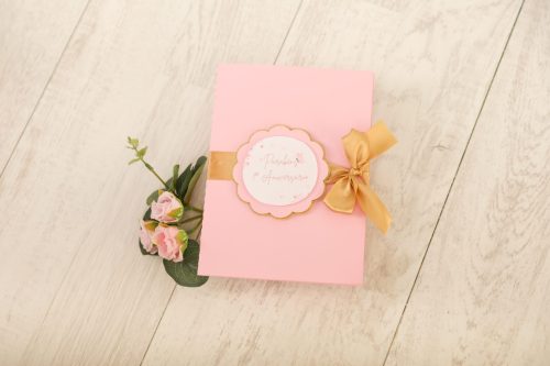 Cadeado rosa com fita dourada e flores, presente delicado e elegante sobre uma superfície de madeira clara.