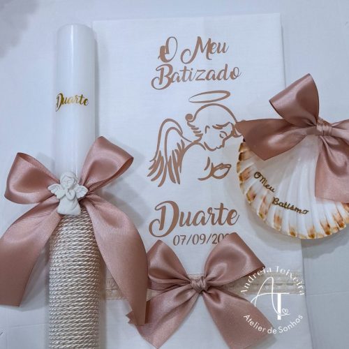 Luminária de vela decorativa com laços de fita rosa, ideal para celebrações de batizado, com uma concha decorativa e uma folha de papel com desenho de anjo e texto em português.