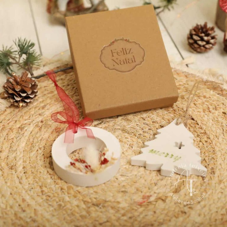 Encarte de presente de Natal com mensagem "Feliz Natal" em caixa de papel kraft, decorado com enfeites natalícios, incluindo uma árvore de madeira e uma guirlanda, sobre uma superfície de palha natura.