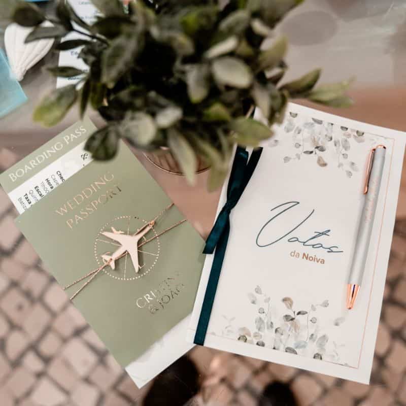 Convite de casamento com tema de passaporte, incluindo um colar de avião e bilhetes de embarque, decorado com plantas e uma caneta dourada.
