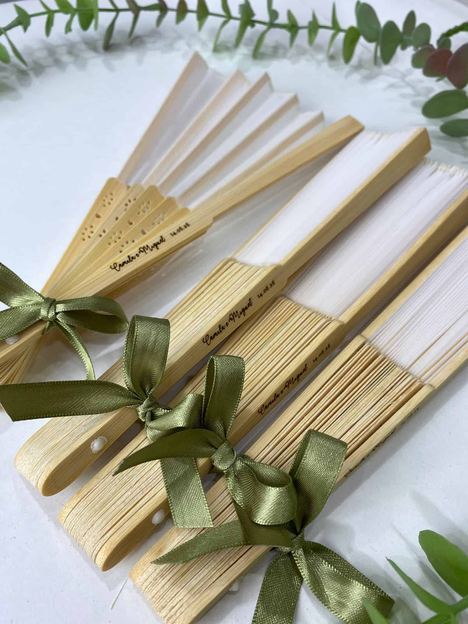 Leque de Madeira Personalizado com Laço 2 Conjunto de leques de papel e madeira amarrados com fitas verdes, decorando uma superfície branca com folhagem ao redor.