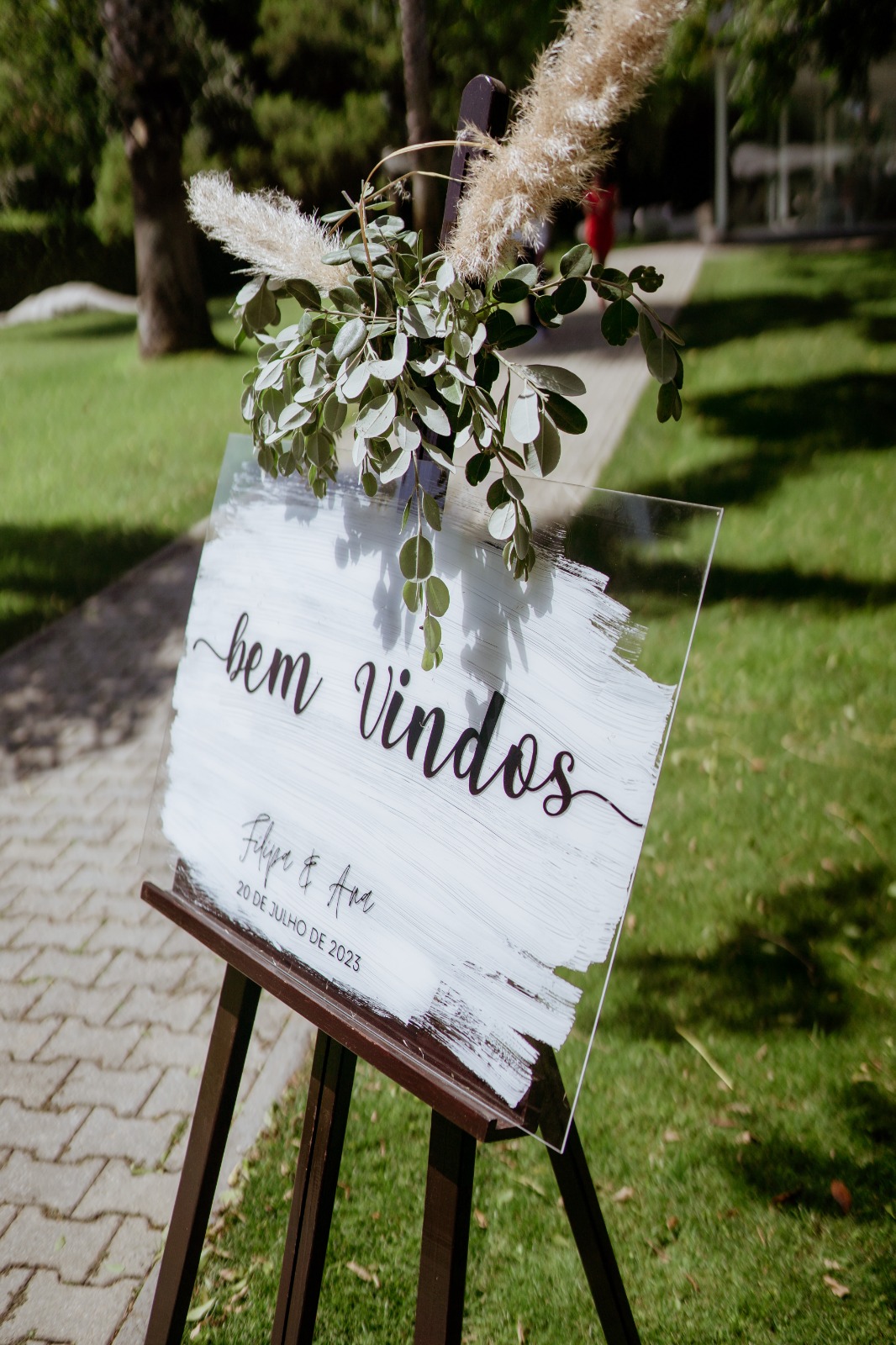Placa acrílica de boas-vindas Coleção Sopro 1 Placa acrílica de boas-vindas da Coleção Sopro para casamento com fundo pincelado a branco assente num cavalete de madeira escura.