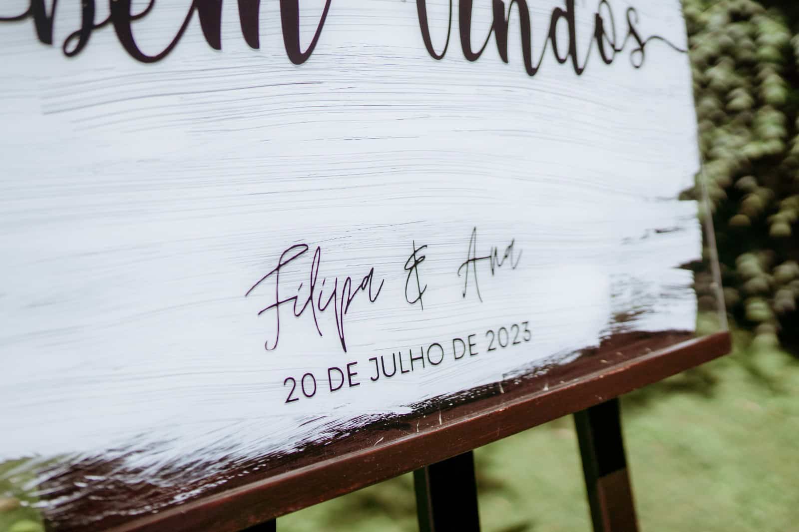 Placa acrílica de boas-vindas Coleção Sopro 2 Placa de madeira com convite de casamento, com nomes Filipe e Ana, e data 20 de julho de 2023, decorada com vegetação ao fundo.