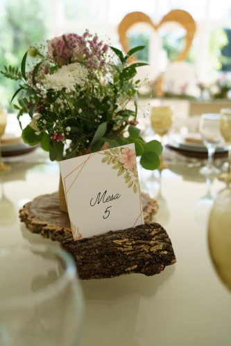 Arranjo floral com flores brancas e roxas em mesa de casamento, com sinal de mesa 5 em suporte de madeira natural.