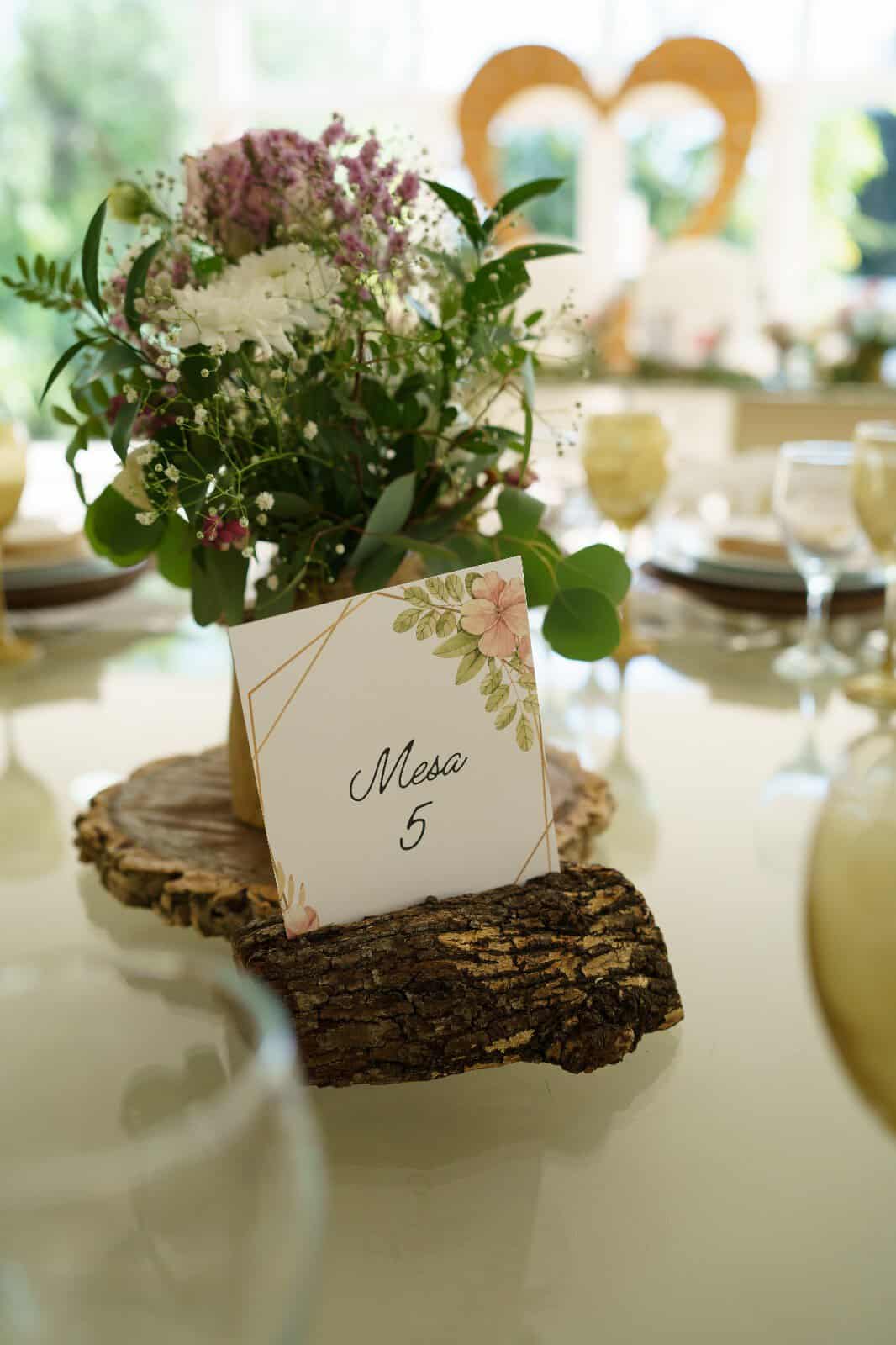Arranjo floral com flores brancas e roxas em mesa de casamento, com sinal de mesa 5 em suporte de madeira natural.