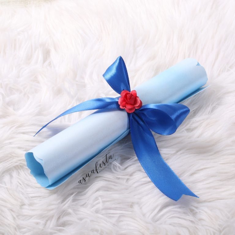 Imagem de uma bandeja de papel azul enrolada, decorada com uma fita de cetim azul e uma pequena flor vermelha no centro, colocada sobre uma superfície de pelúcia branca.