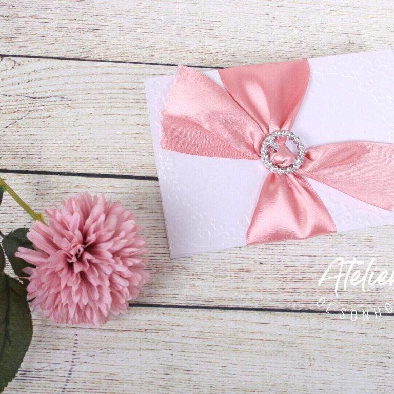 Convite de casamento com laço de cetim rosa, joia e envelope branco sobre mesa de madeira clara.