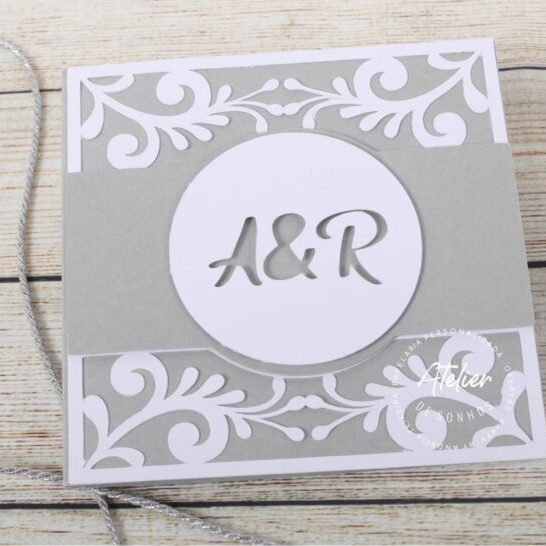 Convite de casamento com design elegante em tons de cinza e branco, apresentando as iniciais A&R em destaque no centro, com detalhes decorativos florais e uma corda para fechamento.