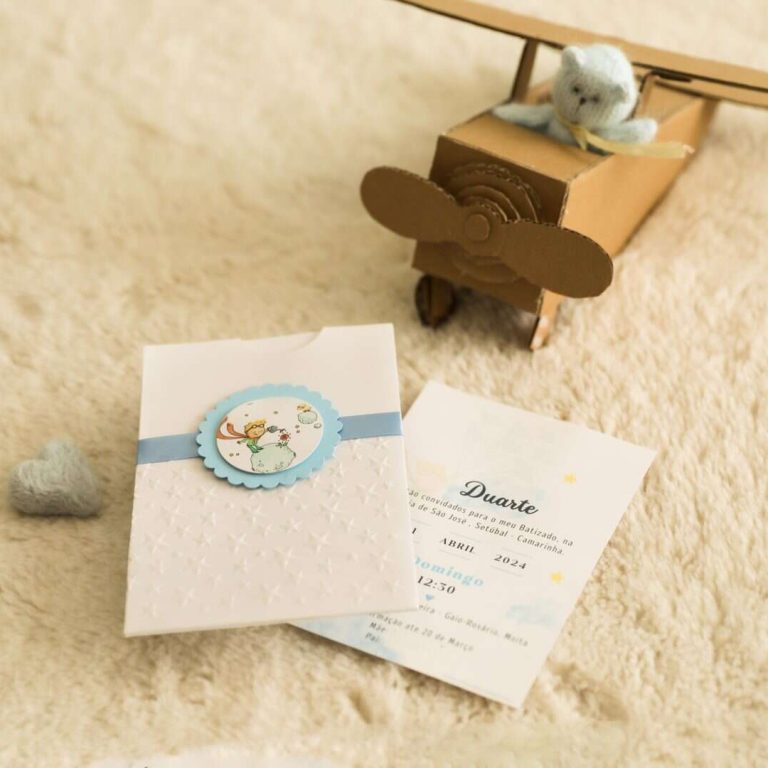 Avião de brinquedo feito de caixas de papelão com um pequeno ursinho de pelúcia no interior, sobre um tapete de pelúcia bege, com convite de batizado ao lado.