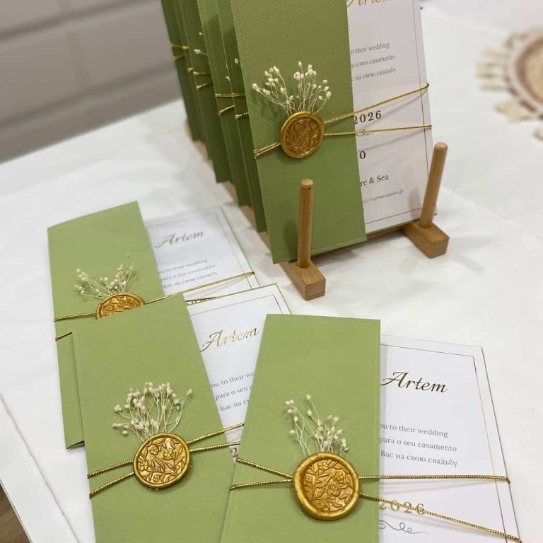Cadeias de convites de casamento com envelopes verdes, detalhes de fita dourada e selo de cera dourado, dispostos em uma mesa branca, prontos para serem entregues aos convidados.