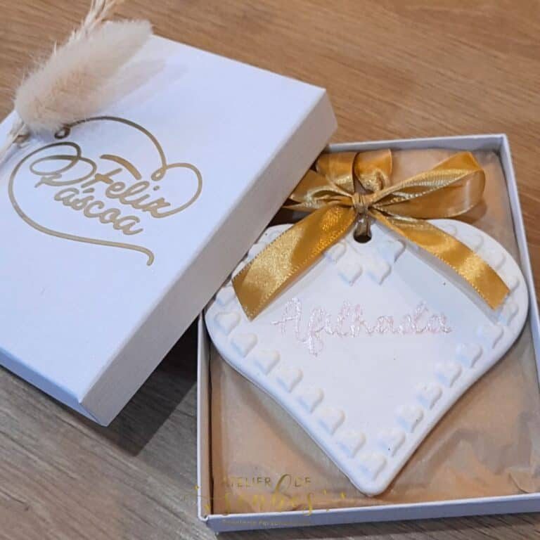 Cadeado de biscuit decorativo com laço dourado e nome personalizado, apresentado numa caixa de presente branca com detalhes dourados e uma pena de pluma, ideal para ocasiões especiais.