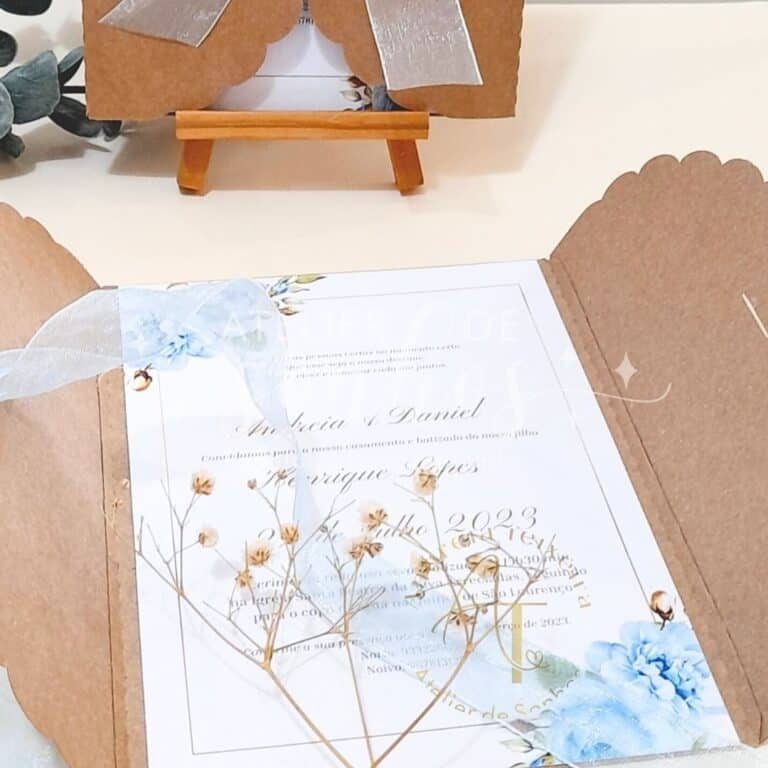 Convite de casamento com detalhes em flores secas e elementos em tons de azul e bege, apresentado em uma caixa de papel kraft.