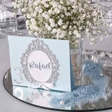 Marcador de mesa da Coleção Aura em cartolina azul clara com numeração, moldura em glitter prateado e laço branco.