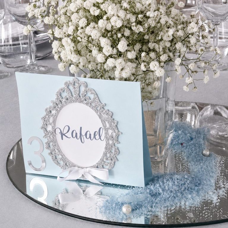 Marcador de mesa da Coleção Aura em cartolina azul clara com numeração, moldura em glitter prateado e laço branco.