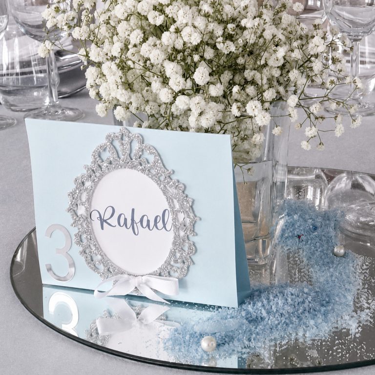 Marcador de mesa da Coleção Aura em cartolina azul clara com numeração, moldura em glitter prateado e laço branco.