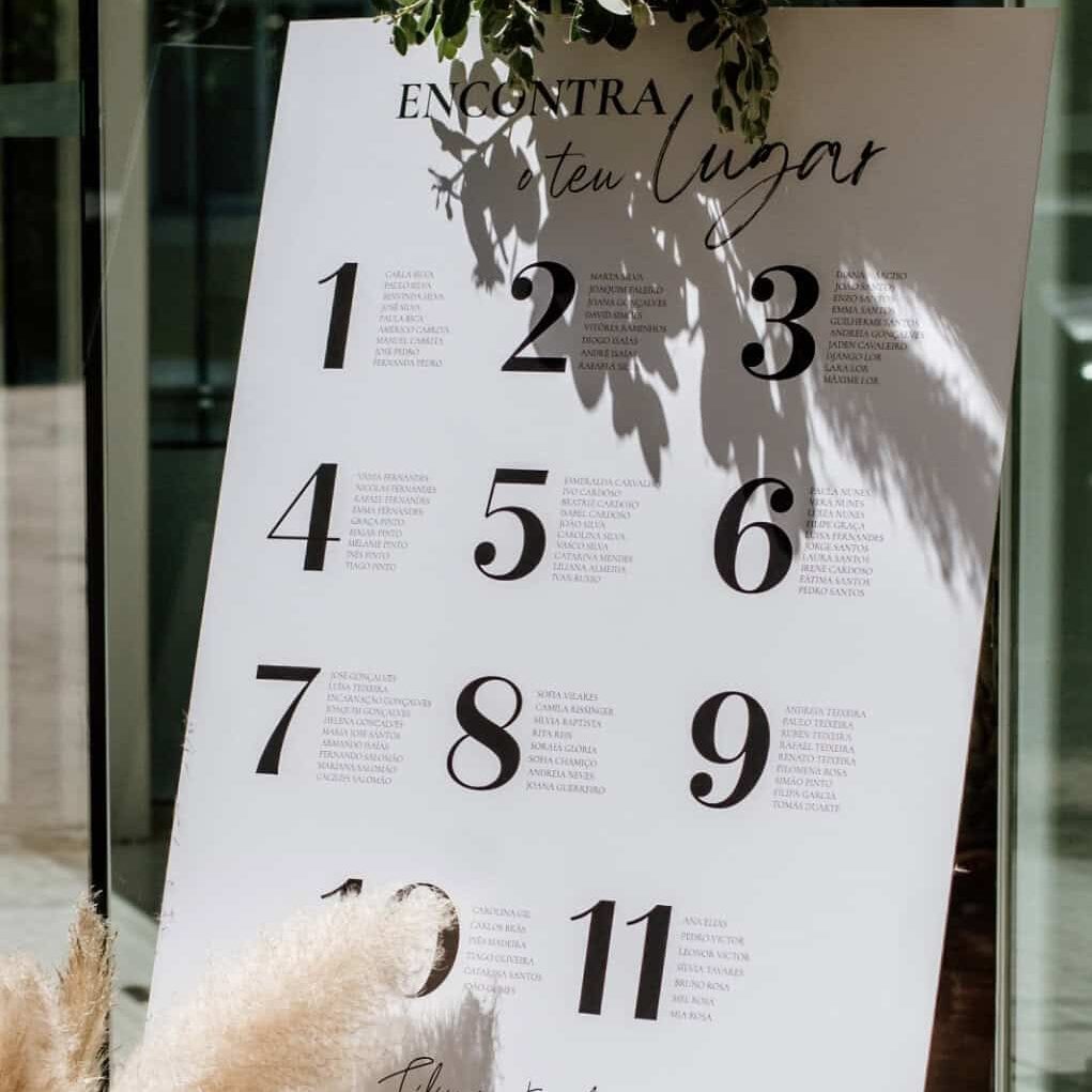Quadro de assentos para casamento com lista numerada e decoração com plantas e flores secas, em ambiente ao ar livre.