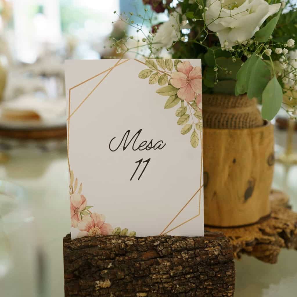Cartaz de mesa com design floral e número 11, decorado com flores brancas e verdes em arranjo rústico de madeira.