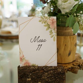 Cartaz de mesa com design floral e número 11, decorado com flores brancas e verdes em arranjo rústico de madeira.