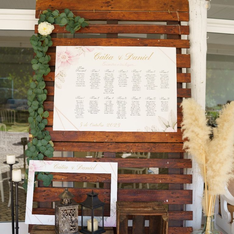 atelier de sonhos artigos de casamento estacionario (8) Cartaz de casamento decorado com folhagem verde e flores brancas, exibindo nomes dos noivos e detalhes do evento, em um cenário ao ar livre com elementos rústicos e iluminação suave.