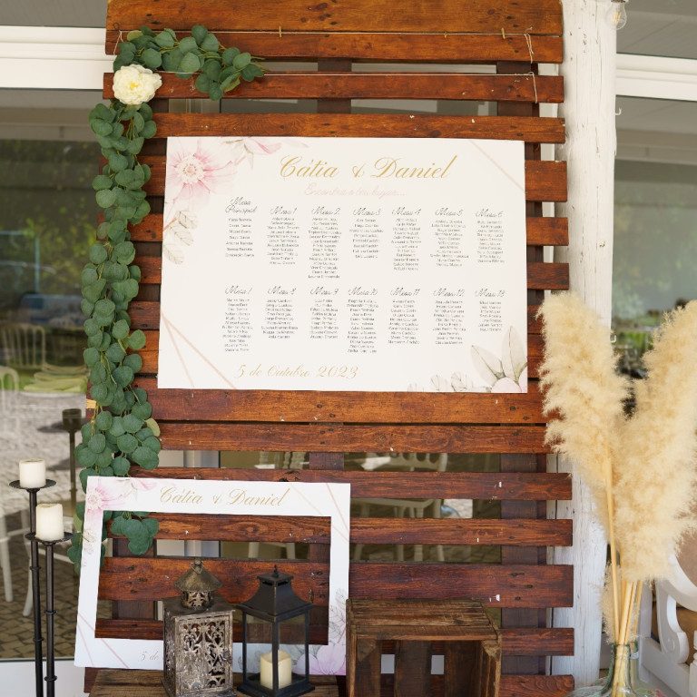 atelier de sonhos artigos de casamento estacionario (8) Cartaz de casamento decorado com folhagem verde e flores brancas, exibindo nomes dos noivos e detalhes do evento, em um cenário ao ar livre com elementos rústicos e iluminação suave.