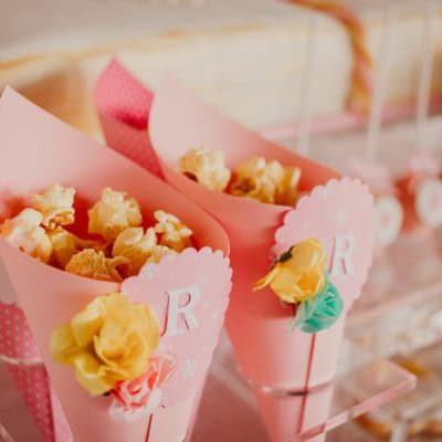 Cone de papel rosa com pipocas decoradas com flores de papel em uma mesa de festa infantil.