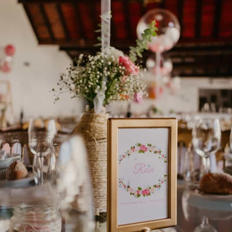 Lata de vidro com flores brancas e rosas, decorando uma mesa de evento com ambiente rústico e elegante, com detalhes de vidro e velas ao fundo.