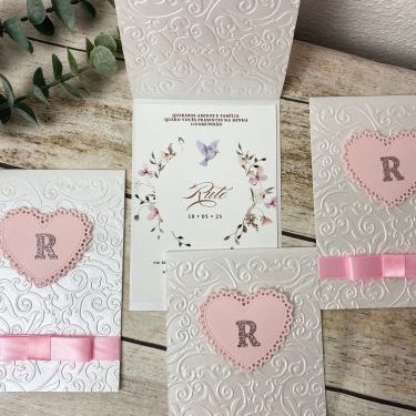 Cadeado de coração rosa com letra "R" em relevo, decorado com fita de cetim rosa, em convite de casamento com fundo branco com relevo delicado.